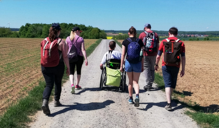 Auf dem Bild sieht man sechs Personen, die einen Feldweg entlanggehen. Eine Person sitzt in einem grünen Rollstuhl, den eine weitere Person schiebt. Alle tragen Rucksäcke und sind sportlich gekleidet. Die Umgebung besteht aus Feldern und Bäumen bei sonnigem Wetter.